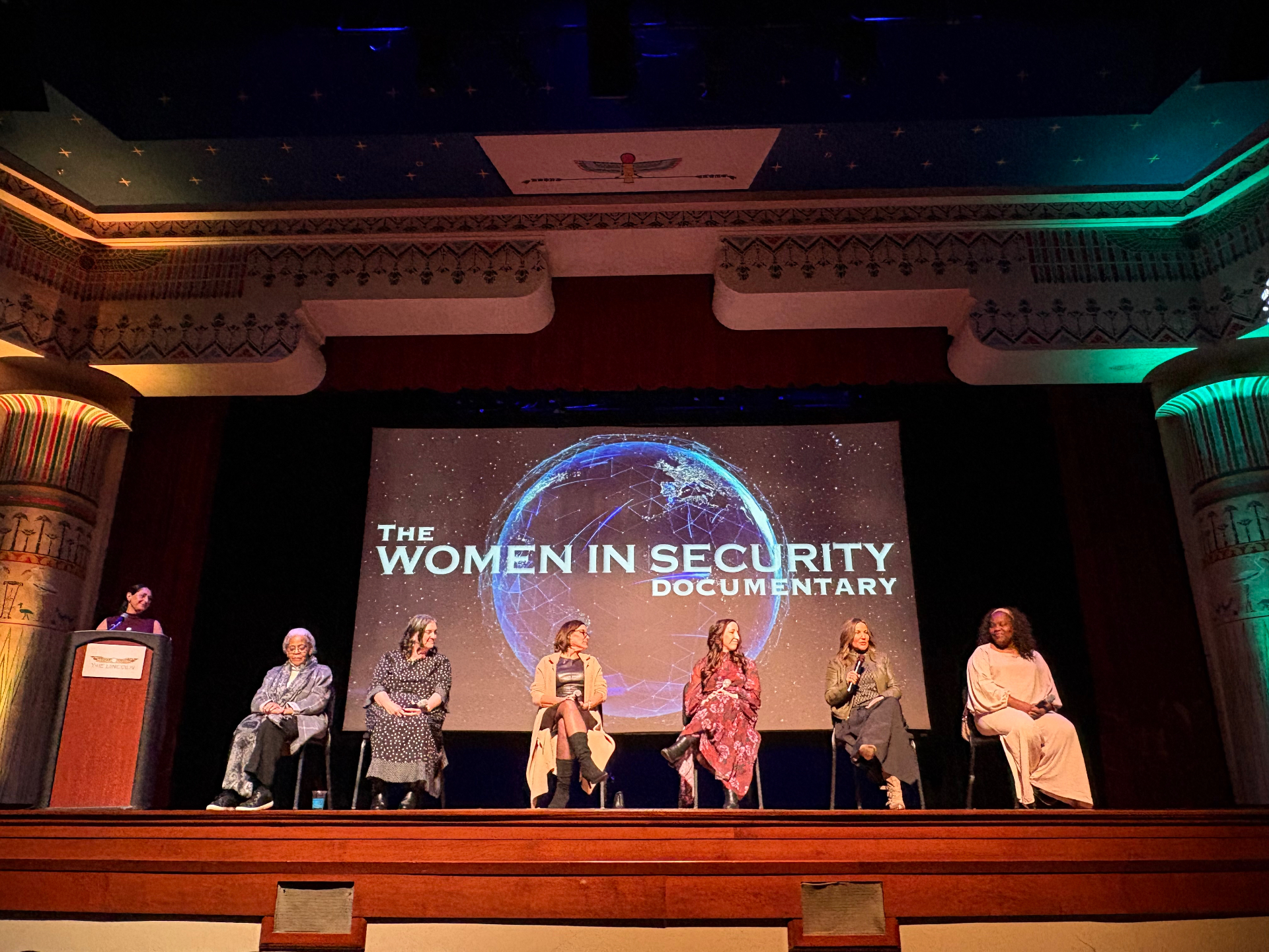 A panel of speakers seated on a historic theater stage beneath the documentary title screen.