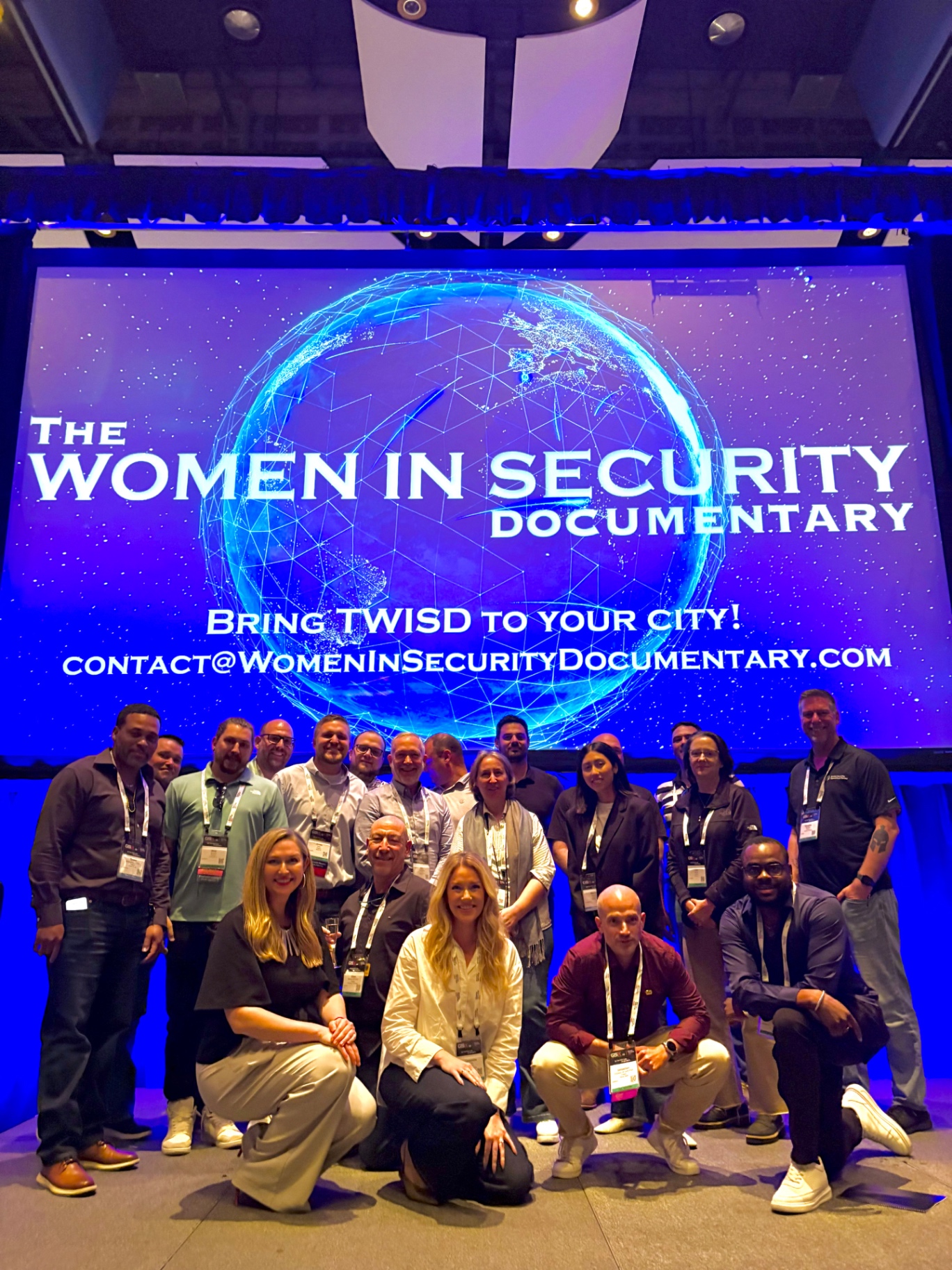 A large group posed on a conference stage in front of the documentary title screen.